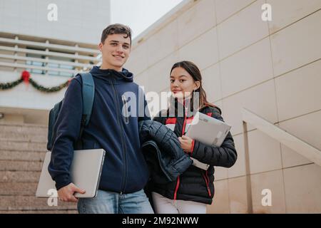 From below positive young students in warm clothes with backpacks and laptops smiling and climbing up stairs while going to university Stock Photo