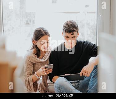Content young friends in casual clothes sitting on windowsill together while surfing internet on gadgets during exam preparation at library Stock Photo