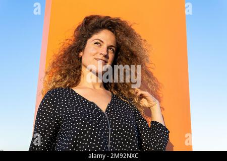 From below smiling young female in polka dot shirt touching brown curly hair while standing against blue sky in the wind Stock Photo