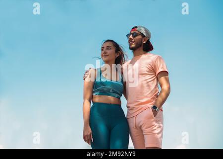 From below young cheerful couple in sportswear embracing looking away standing against on sunny day Stock Photo