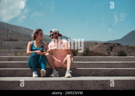 Full body of man in sportswear sunglasses and cap sitting on staircase with girlfriend with mobile phone looking at each other Stock Photo