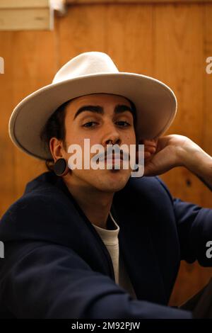Unemotional young Hispanic male model model with dark hair mustache and earplugs in stylish outfit and hat looking at camera against wooden wall Stock Photo