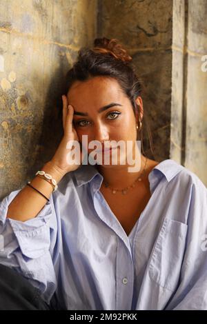 Thoughtful young female tourist with long dark hair in casual outfit sitting leaning head on hand and looking at camera dreamily near aged building Stock Photo