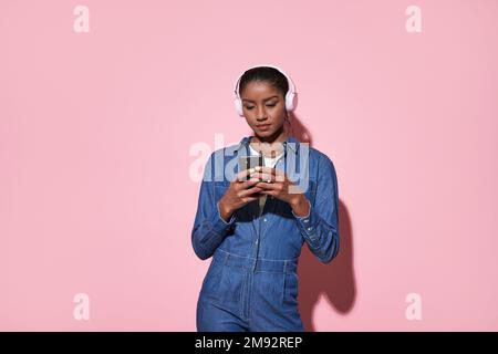 Young focused African American female in denim outfit smiling and listening to music in headphones while standing against pink background with smartph Stock Photo