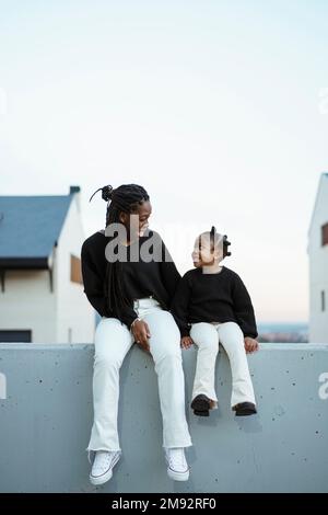 Black woman with Afro braids in casual wear sitting with cheerful girl against residential buildings and cloudless sky having matching outfits looking Stock Photo