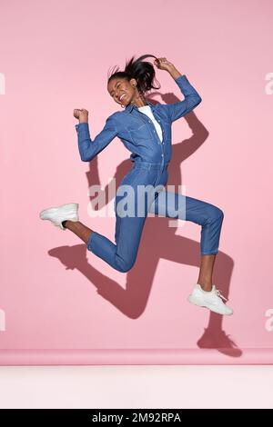 Full body of cheerful young African American female in denim outfit jumping above ground with raised arms and smiling while having fun against pink ba Stock Photo