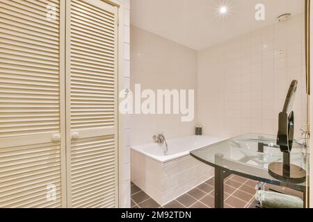 Interior of contemporary bathroom with wooden doors to storage area glass table with sink and bathtub with beige and brown tile Stock Photo