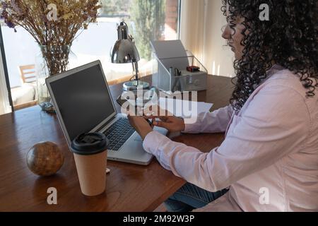 Side view of Hispanic female freelancer with curly hair sitting at table using mobile phone near laptop and takeaway coffee cup at workplace Stock Photo
