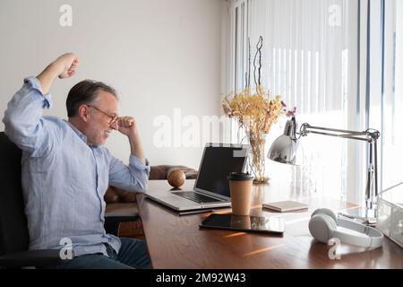 Side view of middle aged excited male freelancer clenching fists and celebrating success while looking at laptop screen during work from home Stock Photo