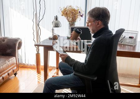 Side view of mature male freelancer in smart casual having coffee in disposable cup while using digital tablet sitting in armchair near table at home Stock Photo