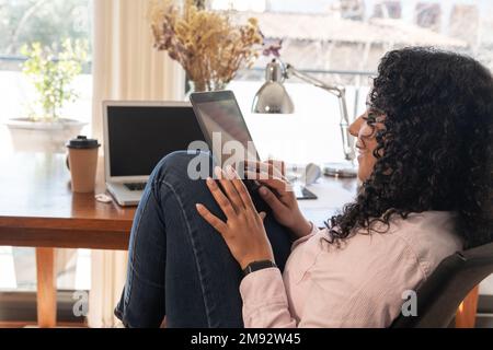 Side view of cropped concentrated female freelancer in smart casual clothes using digital tablet near laptop at table Stock Photo