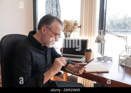 Side view of focused mature male entrepreneur in smart casual sitting in armchair while working on digital tablet at home office Stock Photo