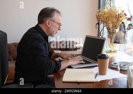 Side view of anonymous mature male freelancer sitting in armchair near table while using laptop and working on project in sunlit living room Stock Photo