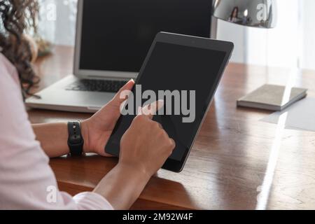 Side view of cropped concentrated unrecognizable female freelancer in smart casual clothes using digital tablet near laptop at table Stock Photo