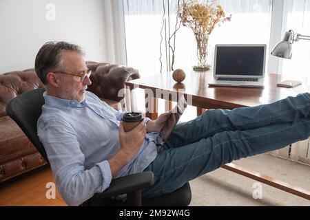 Side view of thoughtful man in casual clothes sitting in armchair with legs on table and drinking coffee while using digital tablet at workplace Stock Photo