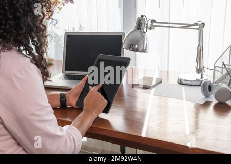 Side view of cropped concentrated unrecognizable female freelancer in smart casual clothes using digital tablet near laptop at table Stock Photo