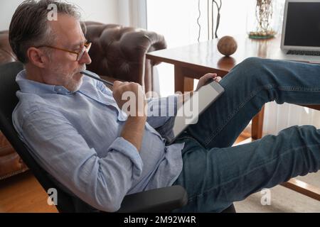 Side view of focused mature male entrepreneur in smart casual sitting in armchair while working on digital tablet at home office Stock Photo