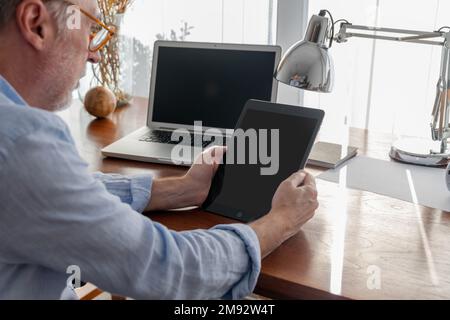 Side view of cropped concentrated mature male freelancer in smart casual clothes using digital tablet near laptop at table Stock Photo