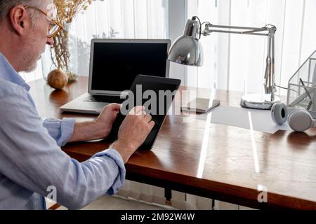 Side view of cropped concentrated mature male freelancer in smart casual clothes using digital tablet near laptop at table Stock Photo