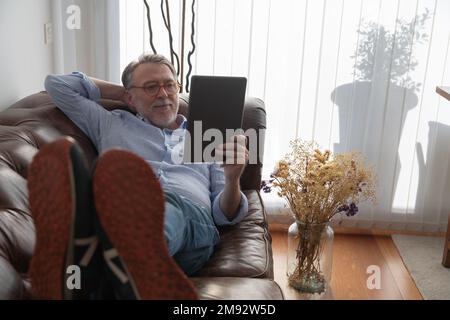 Positive mature male in casual clothes and eyeglasses lying on comfortable sofa while watching video via tablet at home Stock Photo