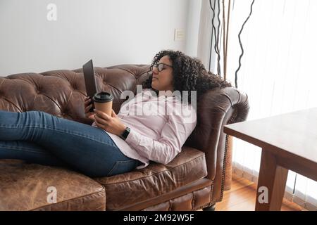 Side view of young ethnic female in casual clothes lying on sofa with cup of hot beverage and using tablet at home Stock Photo
