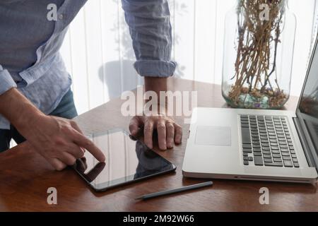 Crop anonymous male freelancer using tablet near laptop on wooden table while working on project at home Stock Photo