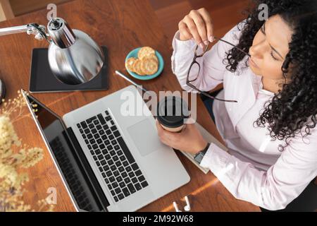 Top view of female freelancer drinking coffee from takeaway cup while working on laptop in wooden table at home office Stock Photo