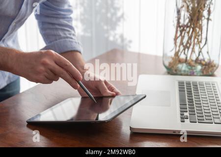 Crop anonymous male freelancer using tablet with stylus pen near laptop on wooden table while working on project at home Stock Photo