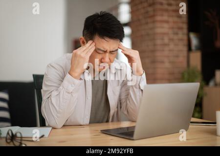Sad mature asian male freelancer working on laptop and suffering from headache, sitting at table at home office Stock Photo
