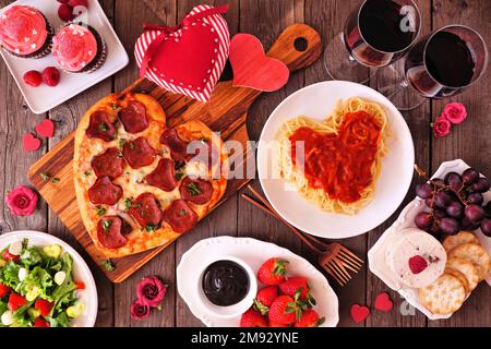 Home cooked Valentines Day dinner. Top view table setting on a dark wood background. Heart shaped pizza, pasta, wine, cheese plate and desserts. Stock Photo