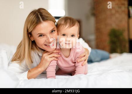 Happy motherhood. Mommy playing with adorable toddler baby crawling on bed, smiling at camera, bedroom interior Stock Photo
