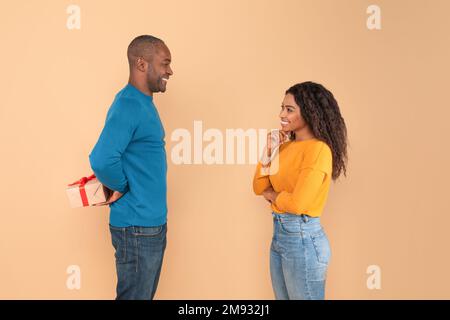 Loving african american man holding wrapped gift box, hiding present for his young wife behind back, side view Stock Photo