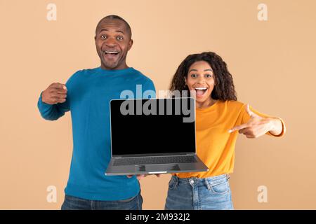 Cool website. Happy black adult couple pointing at laptop with blank screen, man and lady pointing at copy space, mockup Stock Photo