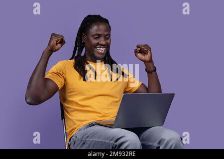 Happy black man sitting on chair, using laptop and gesturing Stock Photo