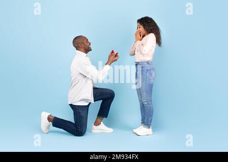 Marry me. Black middle aged man holding giving open box with engagement ring to excited young woman, side view Stock Photo