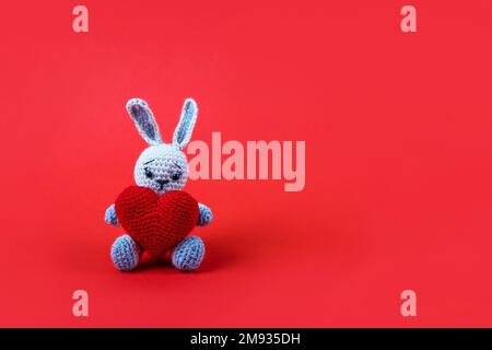 Knitted gray bunny with a crocheted red heart on a red background. Happy Valentine's Day, Mother's Day and birthday greeting card. Stock Photo