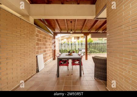 Patio of a detached house with light brown stoneware floors, exposed brick walls, lawned gardens and a porch made of varnished pine logs Stock Photo