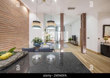 Studio apartment with open kitchen with polished black granite countertop, brick wall, glass dining table and metal pillars Stock Photo