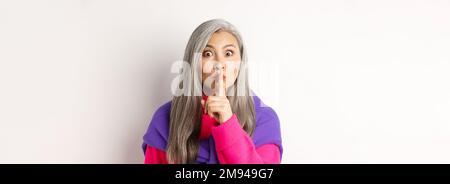 Close-up of stylish senior asian woman in hipster outfit telling hush, shushing at looking at camera, show taboo gesture, standing over white Stock Photo