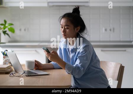Worried upset Asian woman remote worker receiving unpleasant text message while working from home Stock Photo