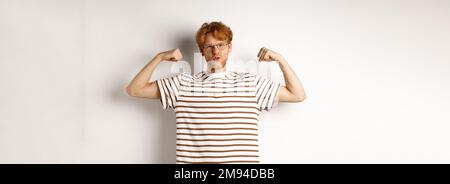 Image of confident and strong redhead man flexing biceps, showing muscles after gym, standing over white background Stock Photo