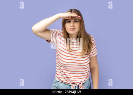 Portrait of attentive beautiful blond woman wearing striped T-shirt standing with hand near forehead and looking far away, looks at distance. Indoor studio shot isolated on purple background. Stock Photo