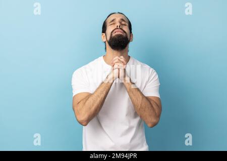 Man wearing white T-shirt looking up holding hands together, praying God, asking for health and support pleading for healing and forgiveness. Indoor studio shot isolated on blue background. Stock Photo