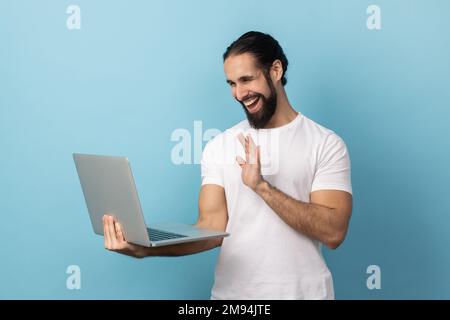Portrait of smiling delighted man with beard wearing white T-shirt having video call, waving hand to camera, saying hello or good bye. Indoor studio shot isolated on blue background. Stock Photo