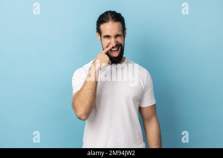 Portrait of crazy handsome man with beard wearing white T-shirt standing and drilling his nose with funny face, winking at camera. Indoor studio shot isolated on blue background. Stock Photo