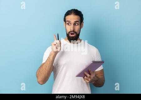 Portrait of man with beard wearing white T-shirt writing creative idea into notebook, taking notes in paper, raised arm, keeps mouth open. Indoor studio shot isolated on blue background. Stock Photo
