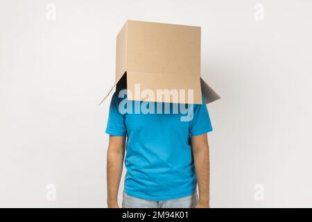Portrait of unknown anonymous man wearing blue T- shirt standing with cardboard box on his head, having fun, hiding his face in carton parcel. Indoor studio shot isolated on gray background. Stock Photo
