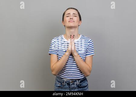 Please, I'm begging. Portrait of upset worried woman wearing striped T-shirt looking up with imploring desperate grimace, praying to god asking for help. Indoor studio shot isolated on gray background Stock Photo