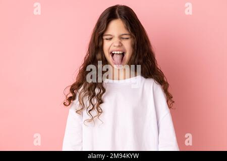 Portrait of childish carefree little girl wearing white T-shirt showing out tongue and closing eyes with naughty disobedient grimace, making face. Indoor studio shot isolated on pink background. Stock Photo