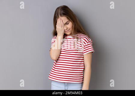 Oh no, I forgot. Portrait of little girl wearing striped T-shirt making facepalm gesture, feeling regret and shame about forgotten event, bad memory. Indoor studio shot isolated on gray background. Stock Photo